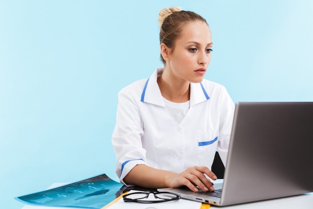 Bela jovem confiante médica vestindo uniforme, sentada no local de trabalho, trabalhando em um computador laptop isolado sobre a parede azul
