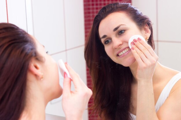Bela jovem de camisa usando creme facial na frente do espelho em seu banheiro