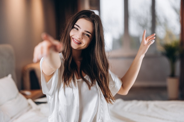 Bela jovem de pijama de seda branca, sorrindo e olhando para a câmera. Bela modelo feminino em um clima alegre está dançando e sorrindo.