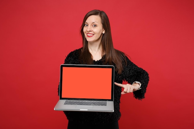 Bela jovem de suéter de pele preta, apontando o dedo indicador no computador laptop pc com tela vazia em branco isolada sobre fundo vermelho. emoções sinceras de pessoas, conceito de estilo de vida. simule o espaço da cópia.