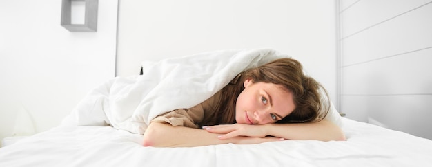 Foto bela jovem deitada em sua cama sob um cobertor olhando para a câmera com uma calma romântica sorrindo