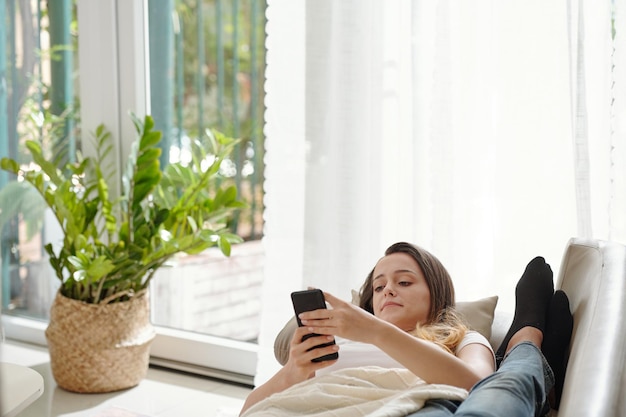 Bela jovem deitada no sofá com o rosto ao lado dos pés do namorado e mensagens de texto de amigos