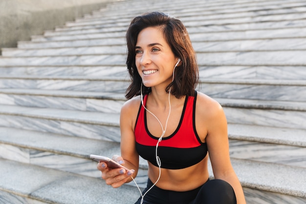 Bela jovem desportista segurando um telefone celular, ouvindo música com fones de ouvido, bebendo água