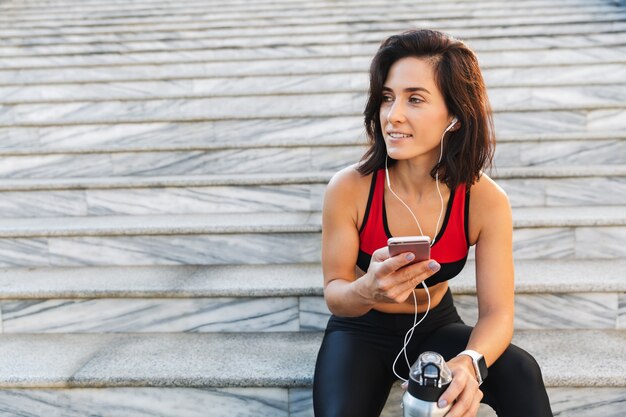 Bela jovem desportista segurando um telefone celular, ouvindo música com fones de ouvido, bebendo água