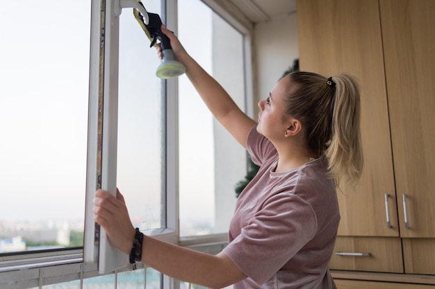 Bela jovem dona de casa trabalhando, lavando janelas por ferramentas profissionais em casa.