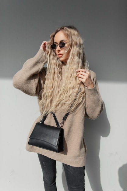 Bela jovem elegante com um penteado loiro encaracolado em um suéter bege da moda com uma bolsa de couro preta coloca óculos de sol vintage e caminha na rua em um dia ensolarado Urban casual girl