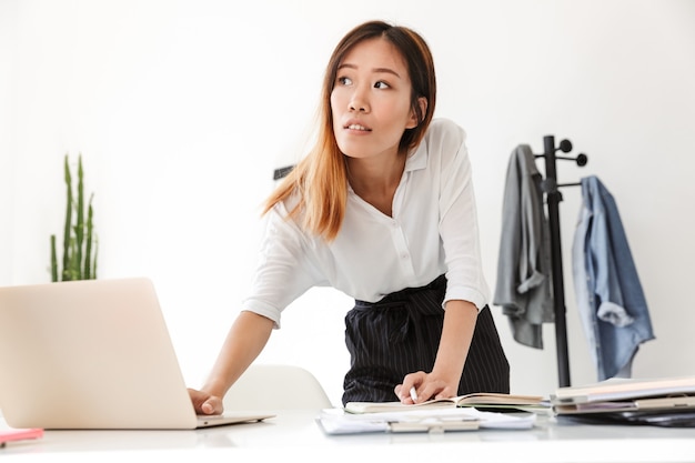 Bela jovem empresária asiática em pé em sua mesa no escritório, trabalhando em um computador laptop