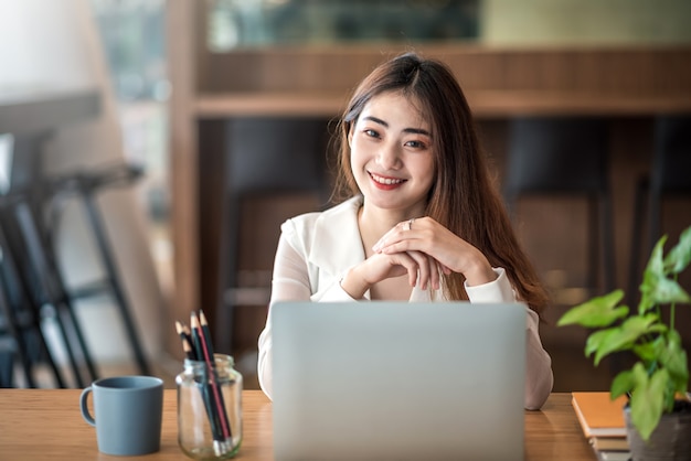 Bela jovem empresária asiática está olhando para a câmera e sorrindo alegremente enquanto está sentado trabalhando no escritório.