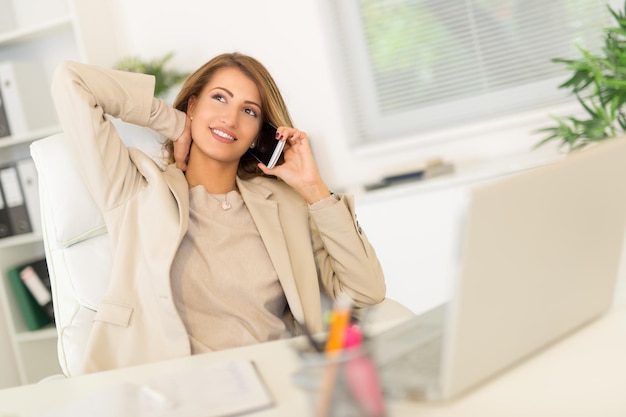 Bela jovem empresária sentada no escritório, relaxando e usando o telefone.