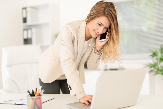 Bela jovem empresária sentada no escritório, usando o telefone e escrevendo no laptop.