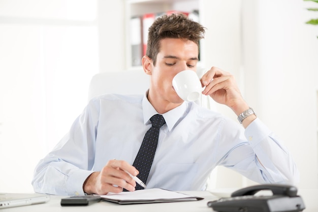 Bela jovem empresário sentado no escritório. Ele está tomando café e trabalhando.