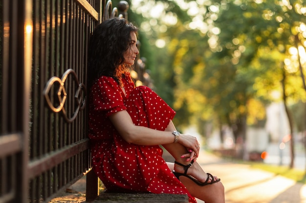 Bela jovem encaracolada com um vestido vermelho vintage e elegantes sapatos de verão sentada na rua