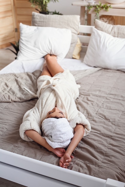 Bela jovem encontra-se na cama vestida com roupão branco