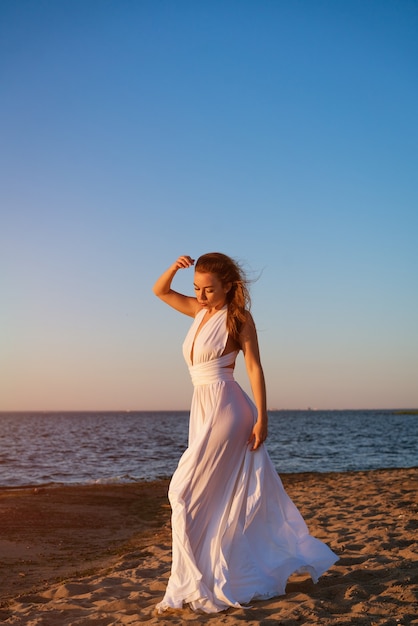 Bela jovem esguia de etnia caucasiana com cabelo comprido fica em um vestido branco no c ...