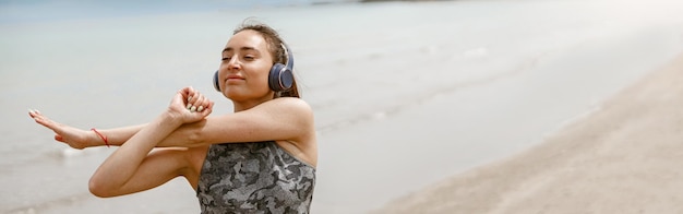 Bela jovem esportiva em fone de ouvido fazendo exercícios de alongamento na praia fundo desfocado