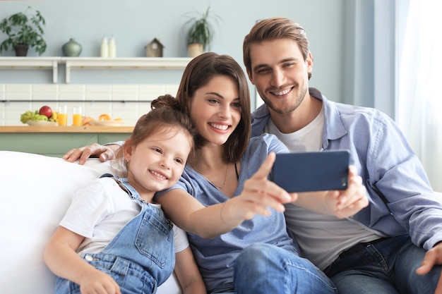 Bela jovem família com criança tomando uma selfie com um smartphone em casa no sofá.