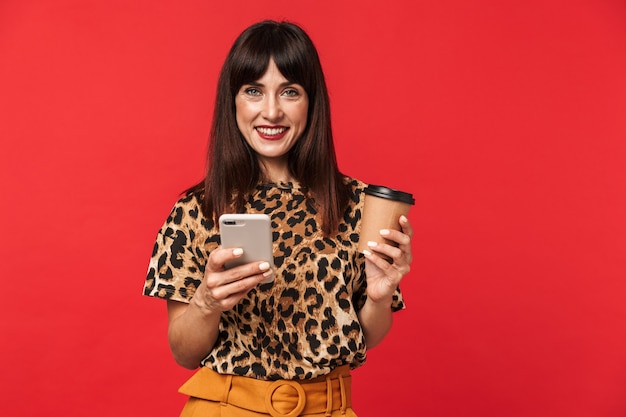 bela jovem feliz vestida com animal impresso camisa posar isolado sobre a parede vermelha, bebendo café usando telefone celular.