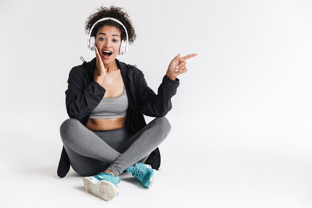 Bela jovem incrível esportes fitness mulher africana ouvindo música em fones de ouvido isolados sobre a parede branca.