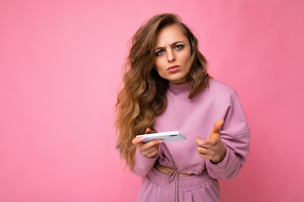 Bela jovem loira com raiva insatisfeita vestindo um capuz rosa isolado sobre um fundo rosa com espaço de cópia segurando e usando o telefone celular, olhando para a câmera