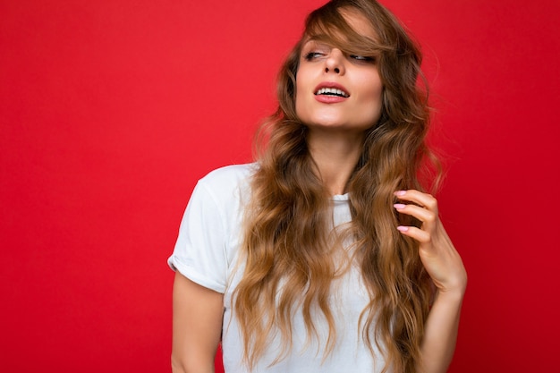 Bela jovem loira encaracolada com expressão sexy, rosto alegre e feliz, vestindo camiseta branca casual isolada sobre fundo vermelho com espaço de cópia.