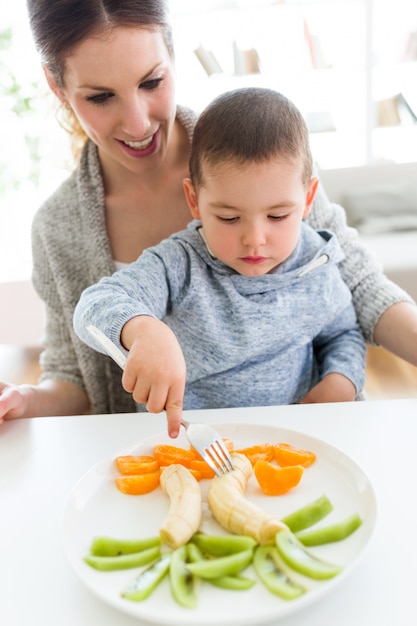 Foto bela jovem mãe e seu filho comendo frutas em casa.