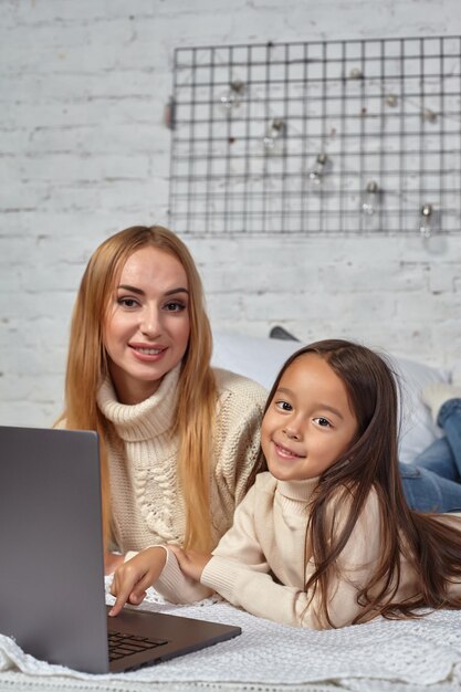 Bela jovem mãe e sua linda filha em suéteres brancos e jeans, deitada na cama em casa la ...