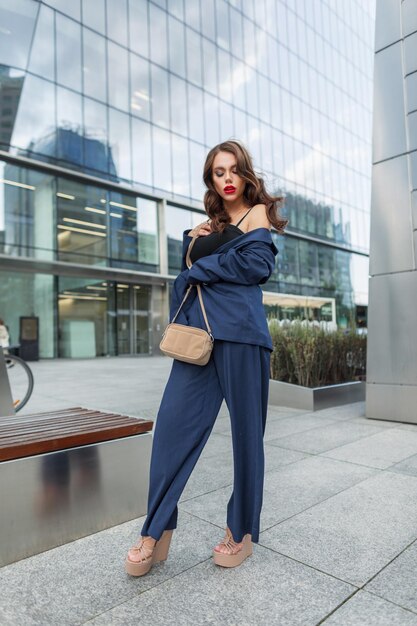 Foto bela jovem moda modelo mulher elegante com penteado em um terno de negócios de moda com blazer calças sapatos com uma bolsa de pé na cidade perto do centro de negócios moderno