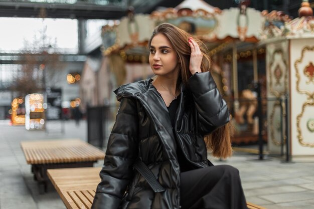 Bela jovem modelo caucasiana em uma jaqueta preta de moda com calças senta-se em um banco na cidade perto de um carrossel vintage
