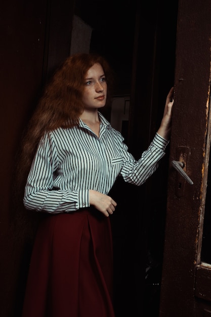 Bela jovem modelo ruiva com camisa stripred e saia vermelha em pé atrás da porta velha