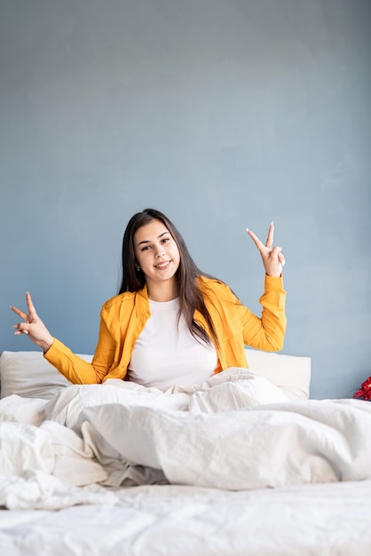 Foto bela jovem morena sentada na cama, mostrando o símbolo da paz com as mãos