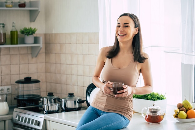 Bela jovem morena sorridente bebe um café maravilhoso e relaxa na cozinha doméstica no fim de semana