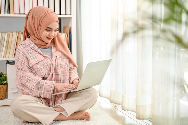 Bela jovem muçulmana asiática usando hijab usando seu laptop na sala de estar