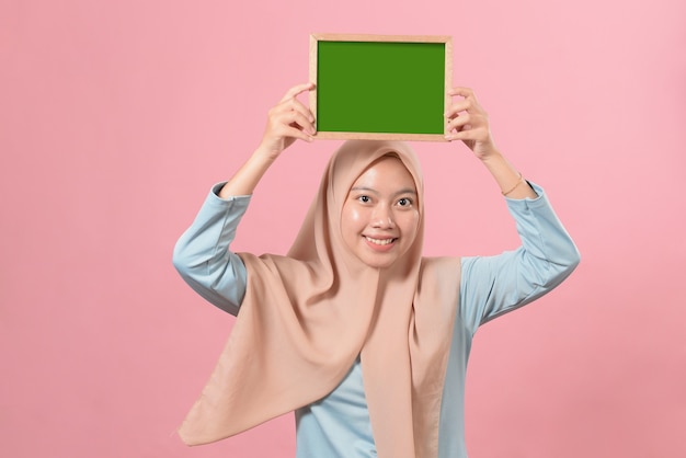 Bela jovem muçulmana segurando e mostrando algo em um quadro-negro sobre fundo rosa