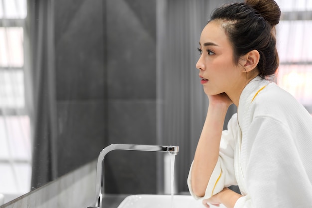 Bela jovem mulher asiática lavando o rosto limpo com água e sorrindo em frente ao espelho no banheiro. Beleza e spa. Pele fresca perfeita