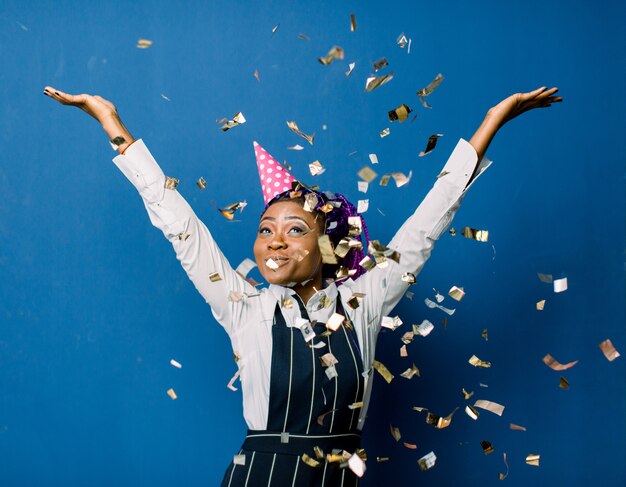 Bela jovem negra comemorando aniversário ou festa de ano novo e chrismas enquanto soprando decorações de confetes para câmera isolada sobre o espaço azul