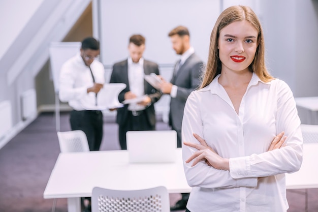Bela jovem profissional sorridente no escritório com as mãos postas e expressão confiante enquanto outros trabalhadores têm uma reunião em segundo plano