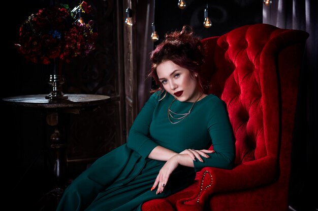 Bela jovem ruiva com maquiagem profissional em vestido verde posando em interior luxuoso no sofá vermelho