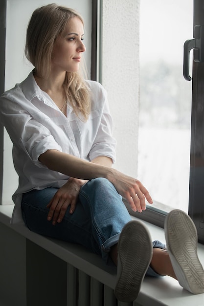 bela jovem senta-se no parapeito da janela em uma camisa branca e jeans e olha pela janela
