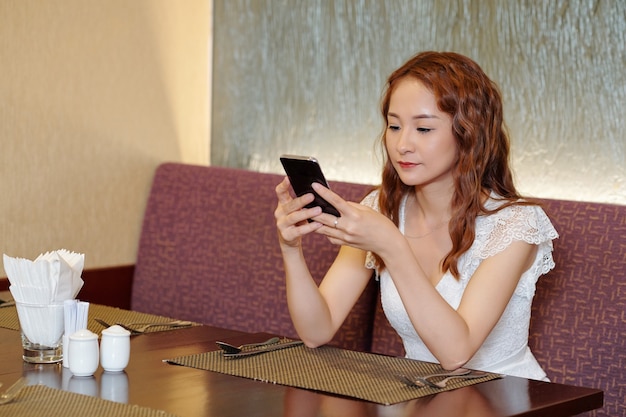 Bela jovem sentada à mesa do restaurante, mandando mensagens de texto para amigos ou acessando as redes sociais