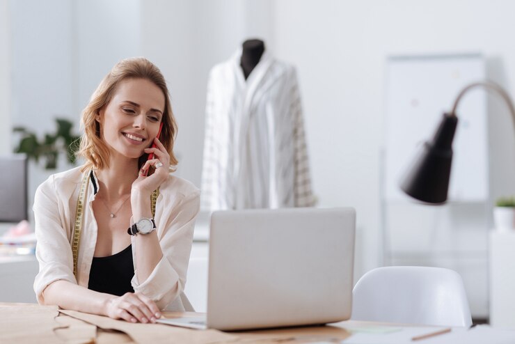 Foto bela jovem sentada à mesa, falando ao telefone e olhando para o laptop com um sorriso encantado.