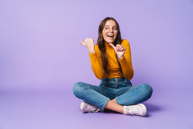 bela jovem sentada com as pernas cruzadas e apontando o dedo para copyspace isolado