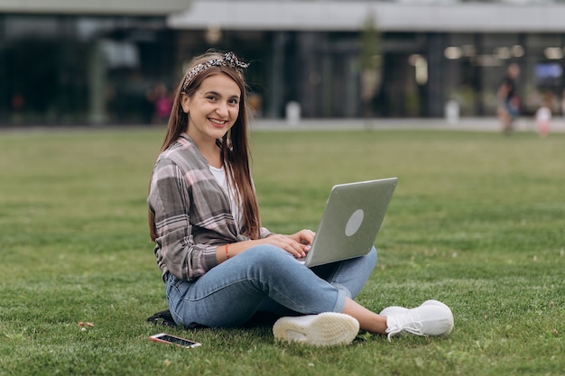 Bela jovem sentada na grama verde no parque com as pernas cruzadas durante o dia de verão enquanto estiver usando o laptop