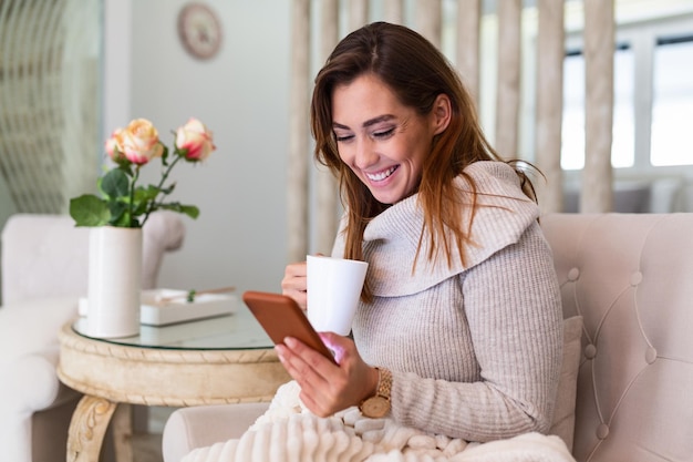 Bela jovem sentada no sofá e apreciando o primeiro café da manhã em um apartamento aconchegante. Mulher sorridente, sentada no sofá enquanto bebe chá quente. Garota feliz relaxando em casa em uma manhã de inverno brilhante.