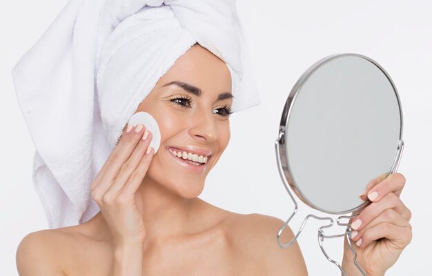 Bela jovem sorridente aplica um tônico no rosto ou remove a maquiagem na frente de um espelho Cosmetologia Garota com uma toalha na cabeça Spa de beleza