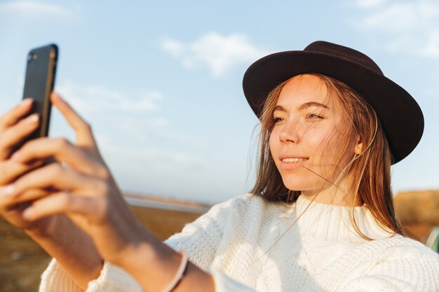 Bela jovem sorridente caminhando ao ar livre na praia durante o pôr do sol, tirando uma selfie