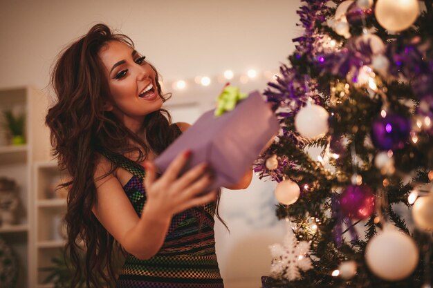 Bela jovem sorridente curtindo o Natal em casa. Ela está segurando um presente ao lado de uma árvore de Natal de decoração violeta e olhando para a câmera.