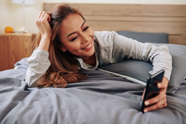 Bela jovem sorridente deitada em uma cama confortável e lendo mensagens de texto no smartphone