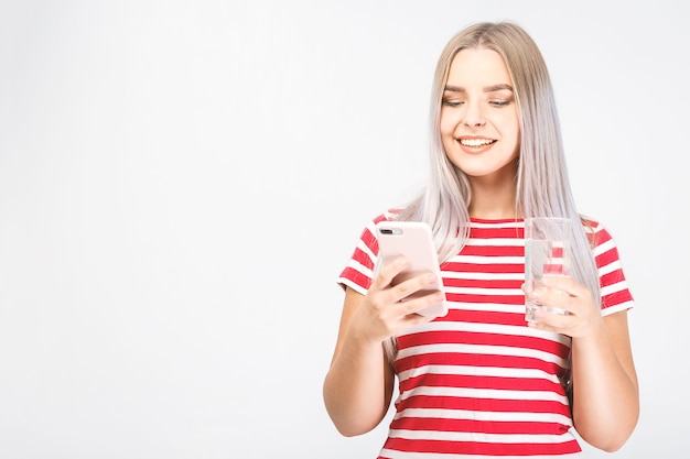 Bela jovem sorridente fica sobre fundo branco e olha para o telefone com um copo de água fria com o celular. isolado, mensagens de texto.