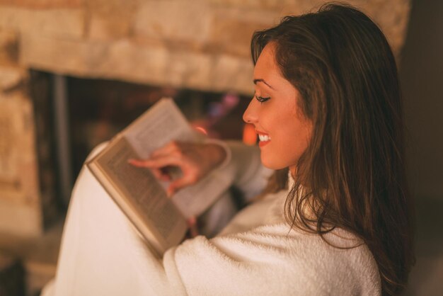 Foto bela jovem sorridente lendo livro e desfrutando junto à lareira.