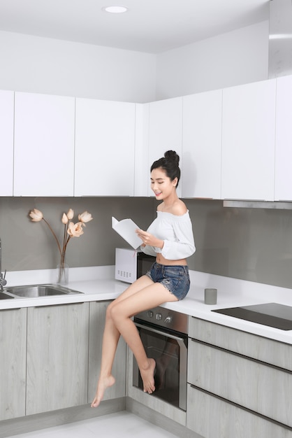 Bela jovem sorridente lendo um livro sentado na cozinha